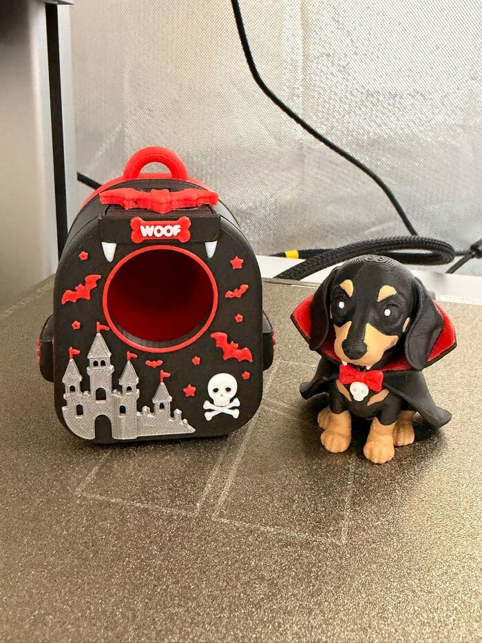 A small 3D printer dog figure in a vampire cape next to a spooky dog house.