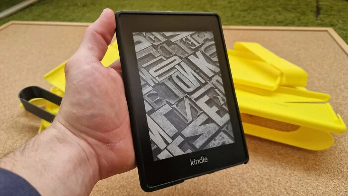 A hand holding a Kindle with yellow 3D printer accessories in the background.