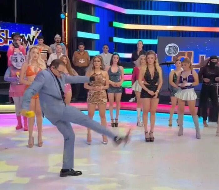 TV show host kicking high on stage in front of a group of female models.