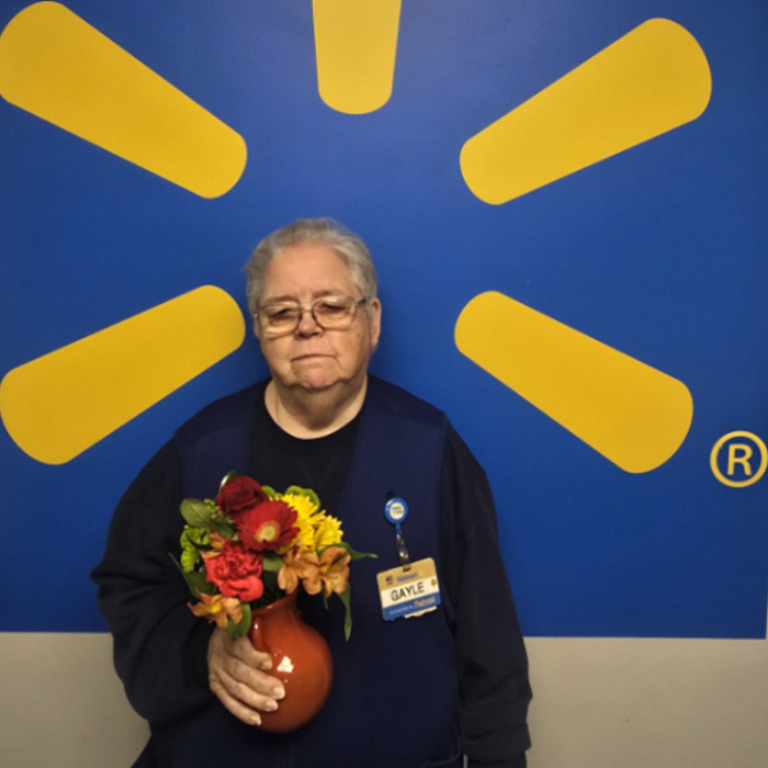 Walmart employee Gayle holding a small vase of flowers in front of a blue background. Walmart employee Gayle holding a small vase of flowers in front of a blue background.