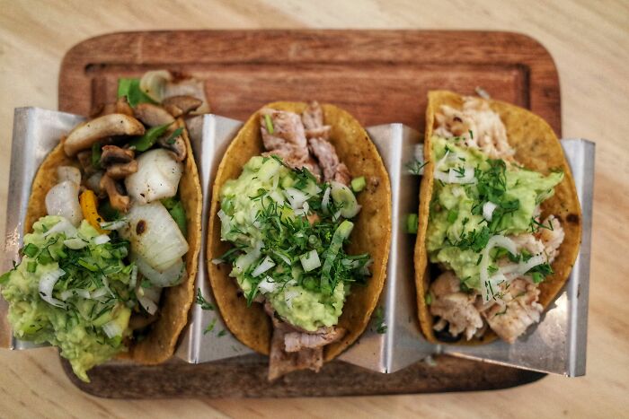 Three tacos topped with guacamole and herbs on a wooden board