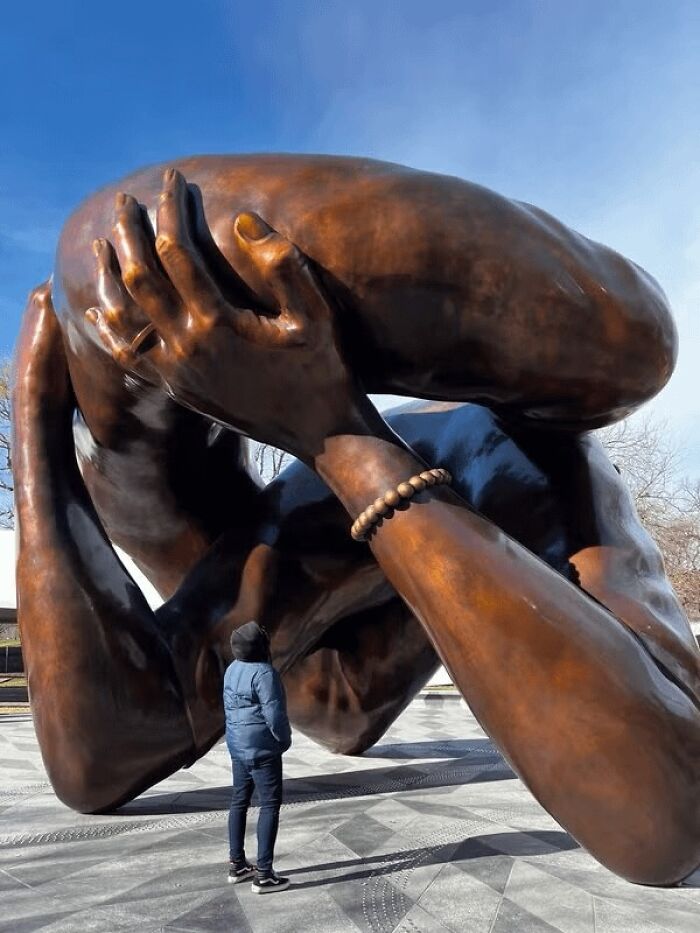 A person looks up at a massive, bizarre public art sculpture representing a giant hug with large bronze arms