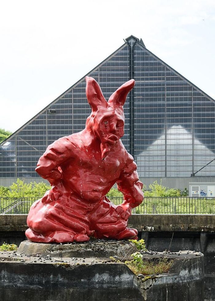 A red, muscular rabbit sculpture, an example of bizarre public art, sitting in front of a modern building.