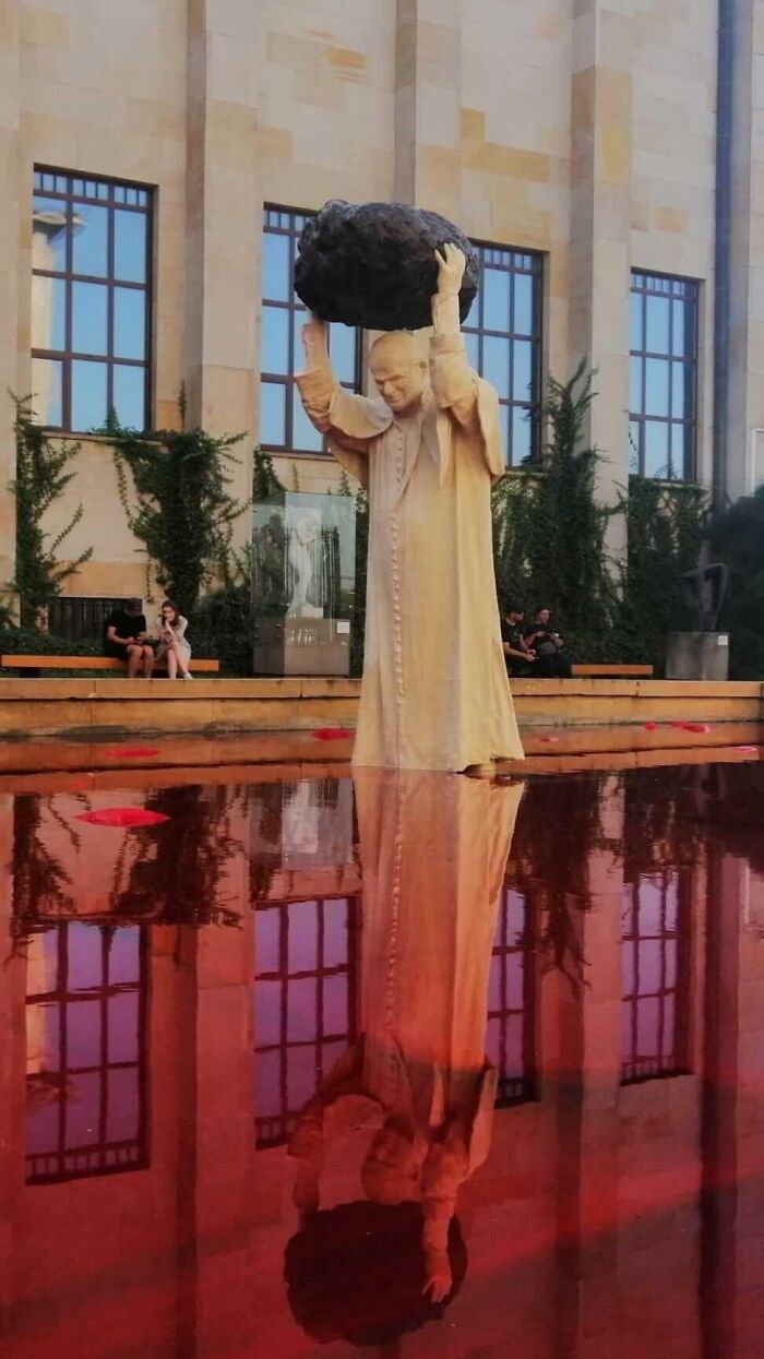 A statue of the Pope lifting a boulder over a red-reflecting pool, a bizarre piece of public art
