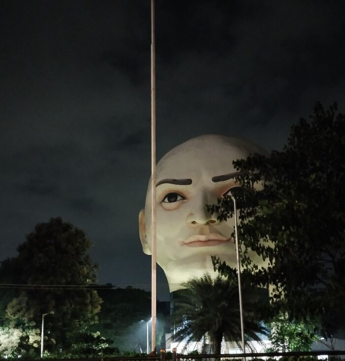 A massive public art sculpture of a realistic human head peeking out behind trees at night.
