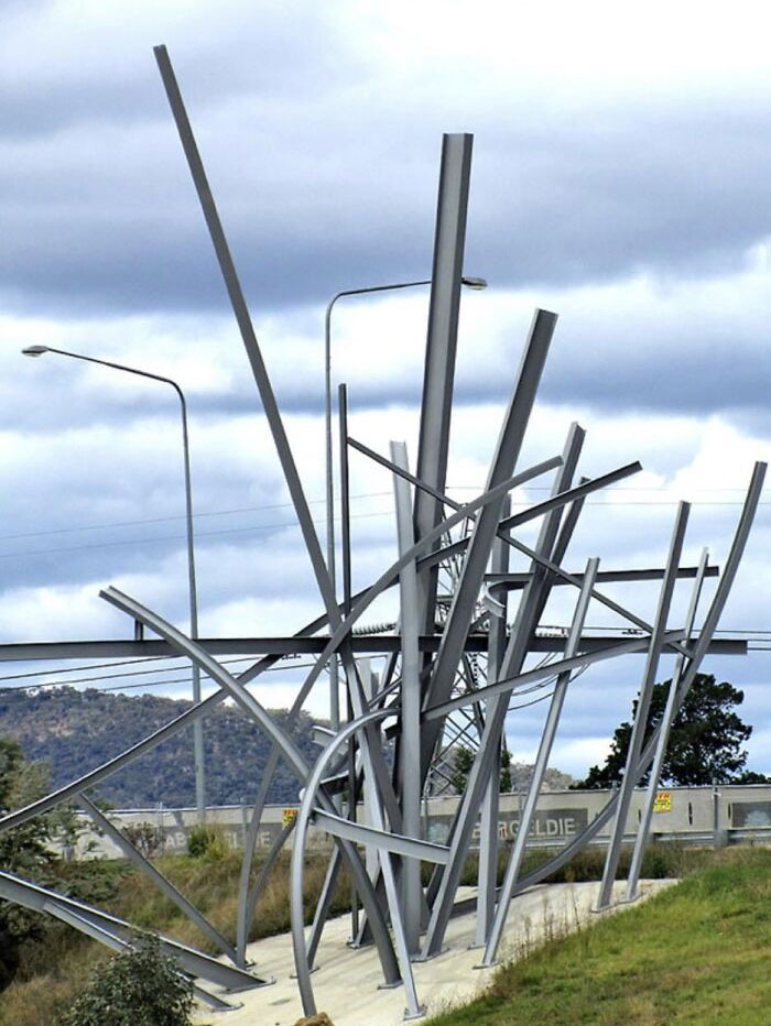An abstract public art sculpture made of haphazardly arranged silver metal beams under a cloudy sky.