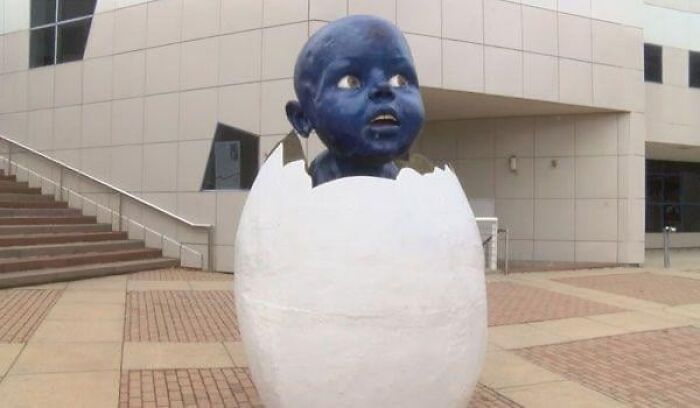 A creepy public art statue of a blue baby hatching from a white egg shell