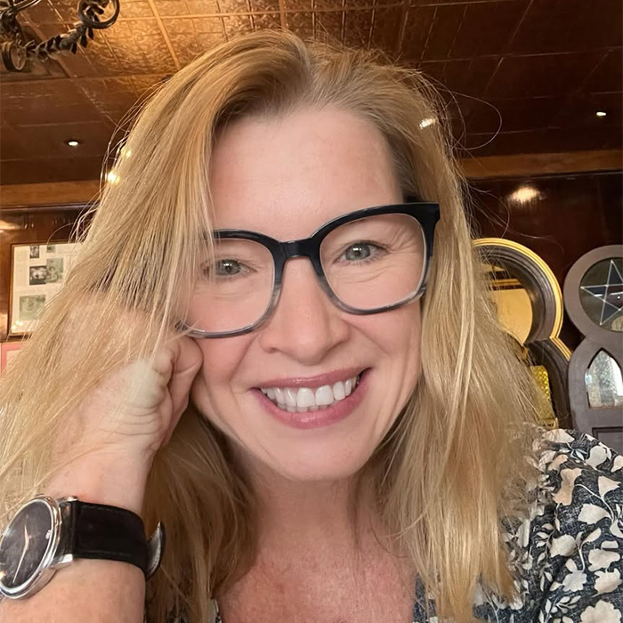 A smiling blonde woman wearing glasses resting her chin on her hand, sharing her story after cancer diagnoses.