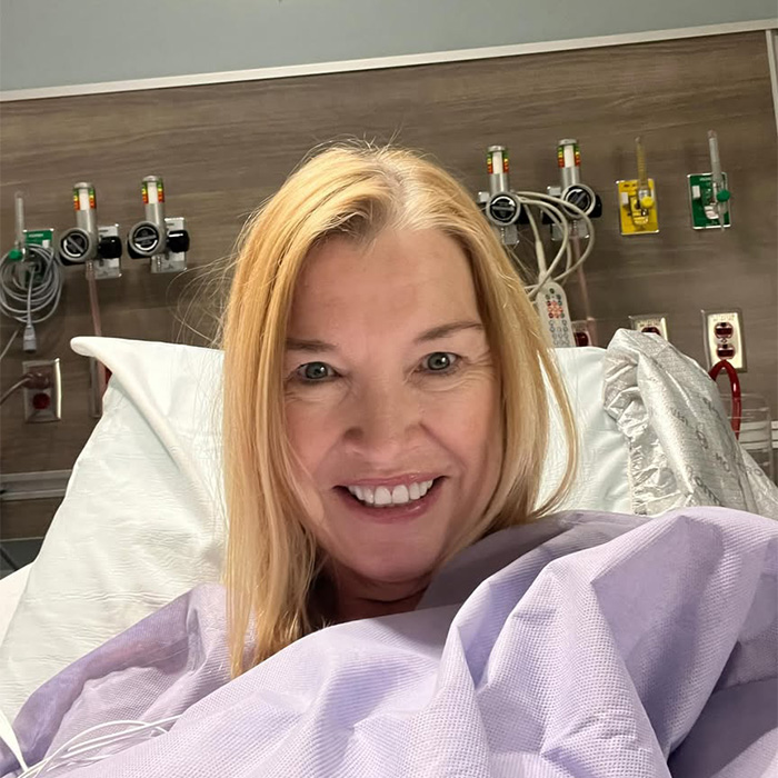 A blonde woman smiling in a hospital bed, facing treatment following medical news about cancer diagnoses.