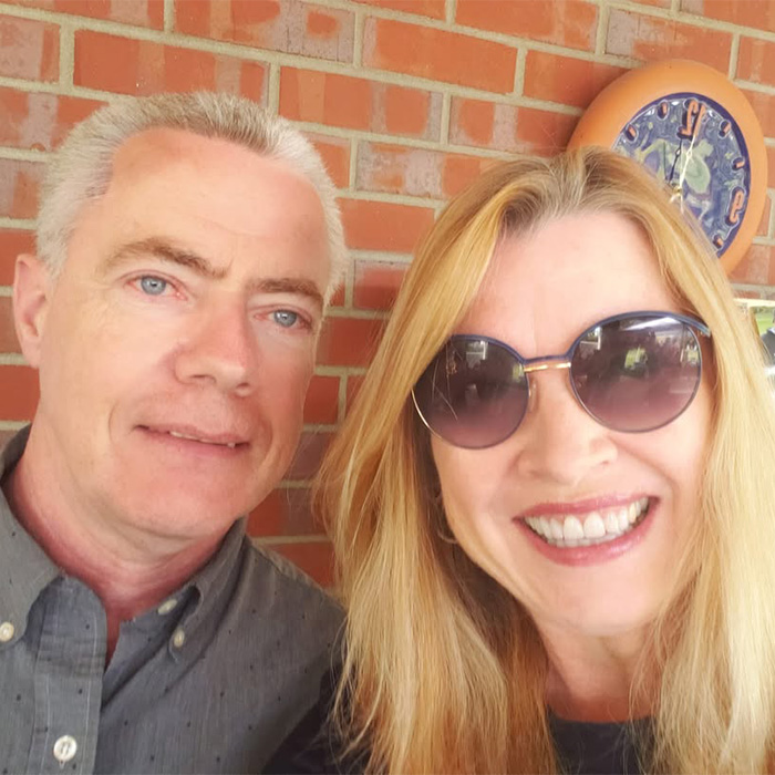 A blonde woman in sunglasses smiling next to a man with short gray hair, reflecting on her husband's behavior.