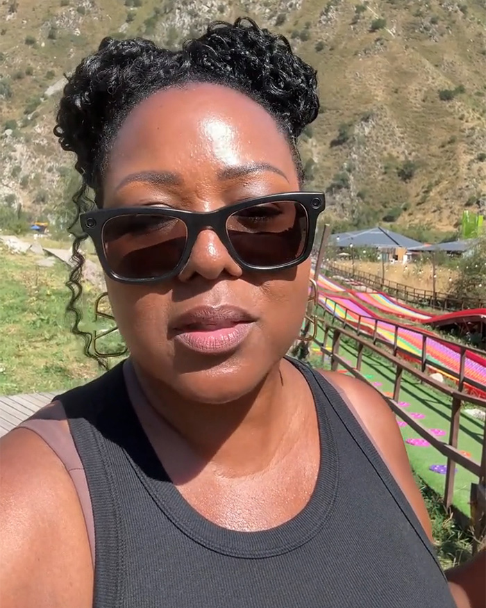 Woman wearing black tank top and sunglasses outdoors with hillside and colorful slides