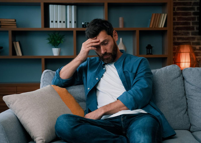 Stressed man sitting on a couch holding his head in distress, reflecting on a fiancé’s lie before getting married. Stressed man sitting on a couch holding his head in distress, reflecting on a fiancé’s lie before getting married.