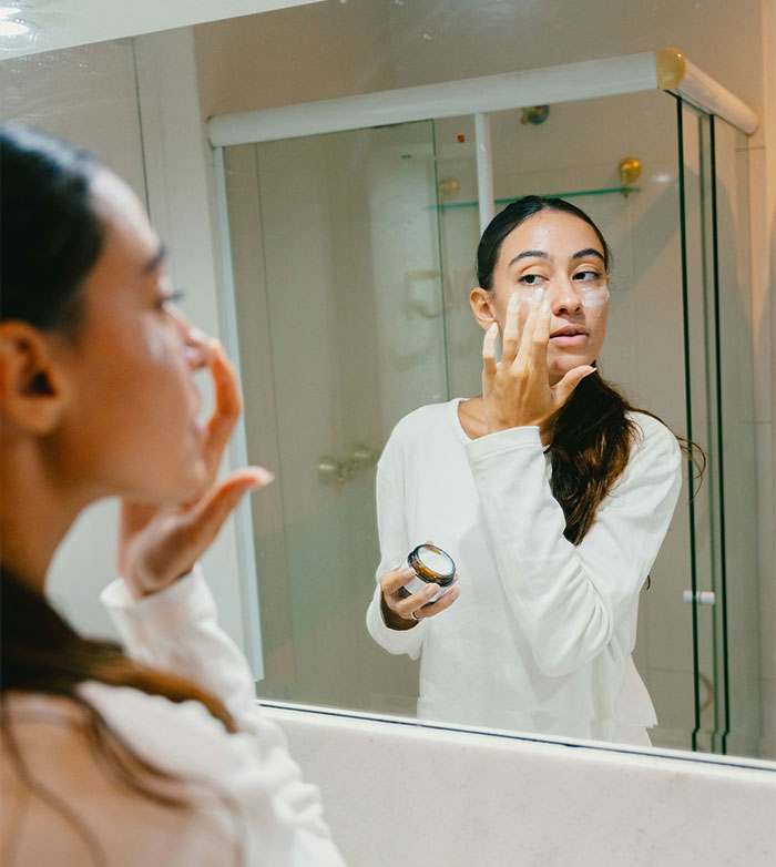 Woman applying soothing cream to her face while looking in the mirror after lip filler treatment Woman applying soothing cream to her face while looking in the mirror after lip filler treatment