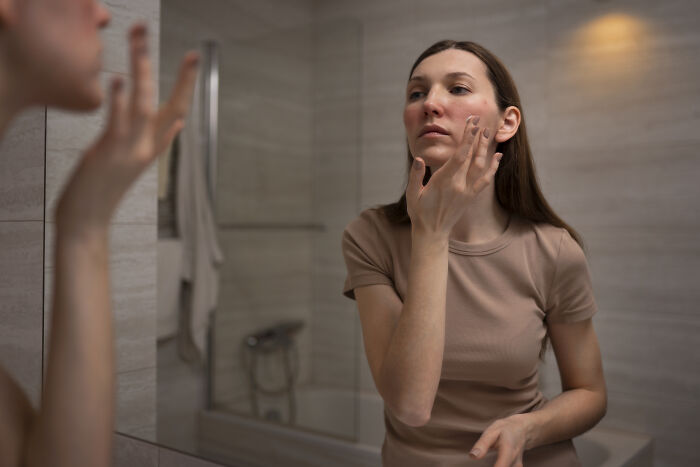 Woman applying face cream while looking in a bathroom mirror, treating Gen Z health problems like acne.