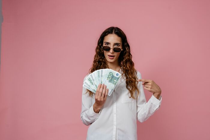 A woman in a white shirt and sunglasses holding a fan of euro bills, showing people behaving like they are owed everything
