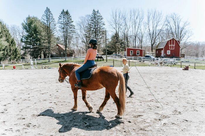 A young woman riding a brown horse in an outdoor arena while a trainer guides her, depicting entitled behavior themes