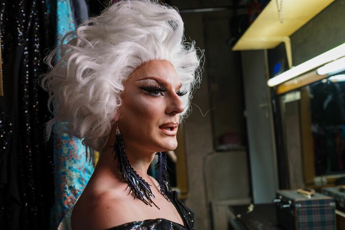 Person in drag makeup and white wig looking in dressing room mirror