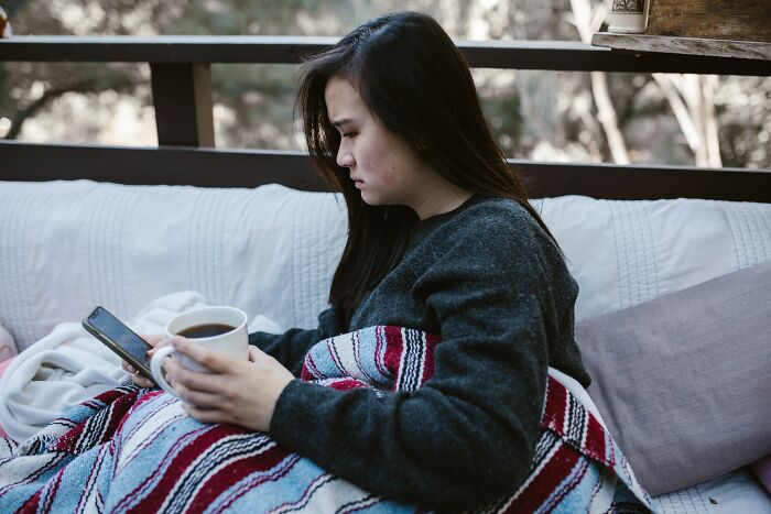 Woman wrapped in blanket looking at phone with coffee cup