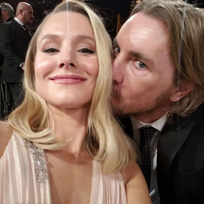 Kristen Bell smiling next to Dax Shepard who kisses her cheek during an event.